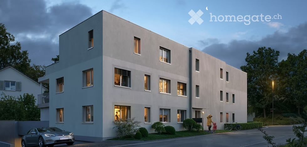 Modern two-story residential building with clean lines and large windows, illuminated at dusk. The building features a flat roof and is surrounded by small landscaped areas with shrubs and trees. A car is parked in front, and a person is walking near the