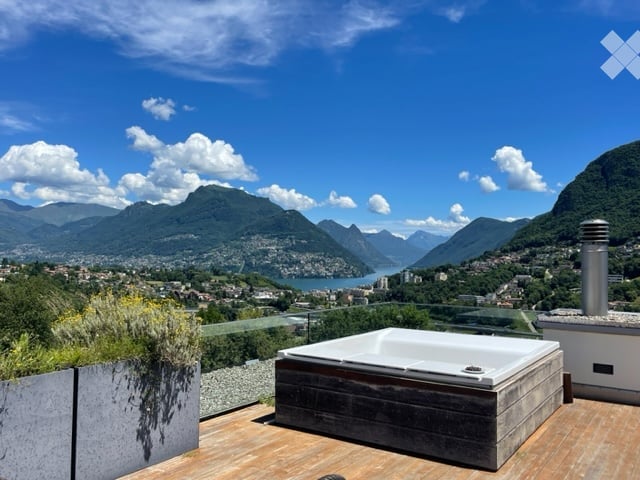 A modern rooftop terrace with a hot tub, wooden decking, and expansive views of a lake and surrounding mountains.