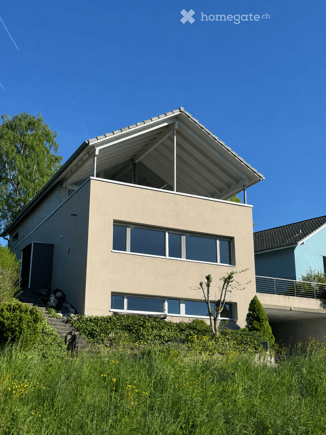 Modern two-story house with a flat roof and large windows, situated on a raised ground floor with a grassy slope leading up to it.