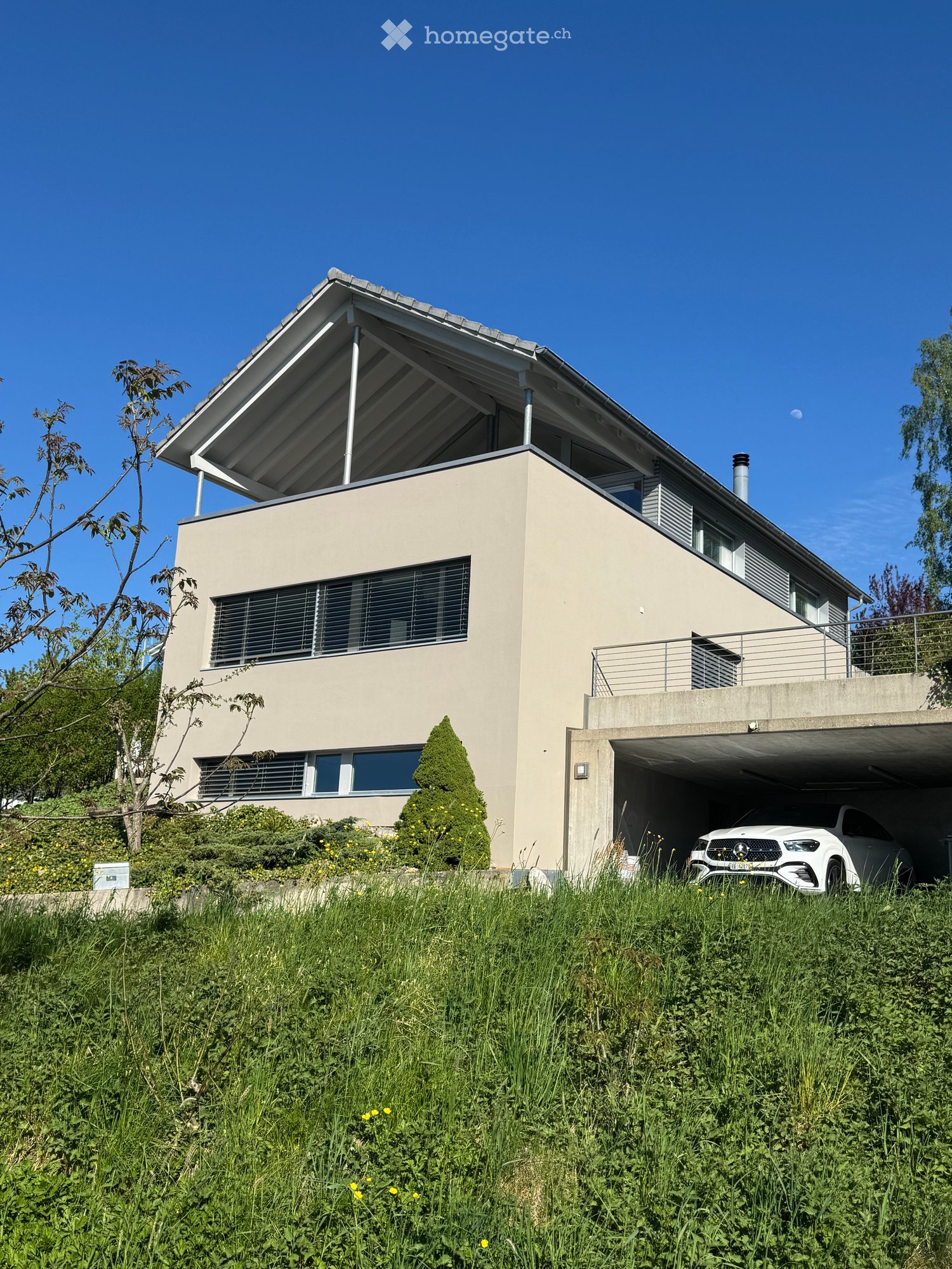 Modern two-story house with a flat roof, large windows, and a covered garage. The house is surrounded by greenery and has a raised ground floor.