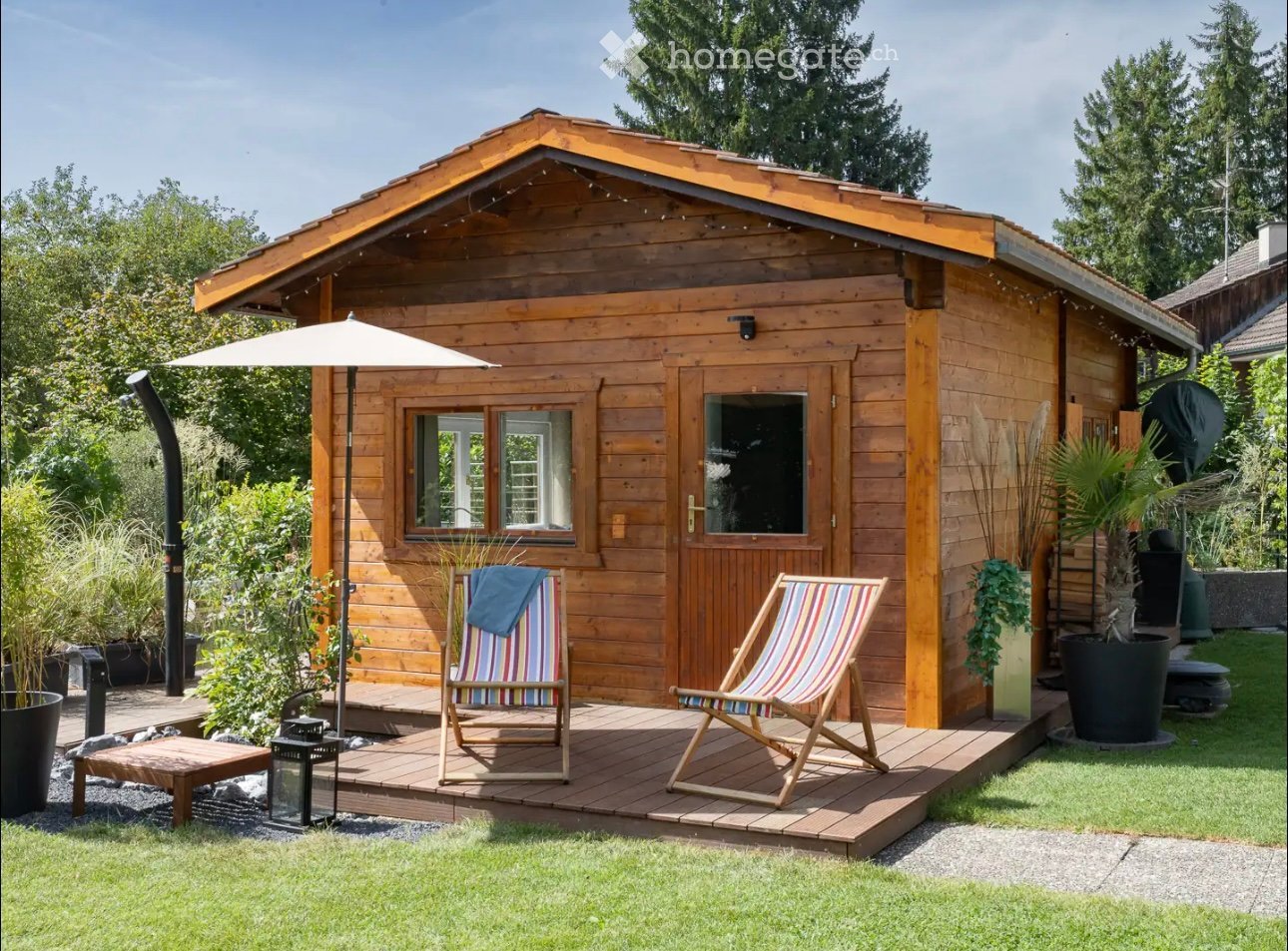A wooden cabin with a raised deck, umbrella, two striped chairs, small table, and lantern. The deck is surrounded by potted plants and greenery.