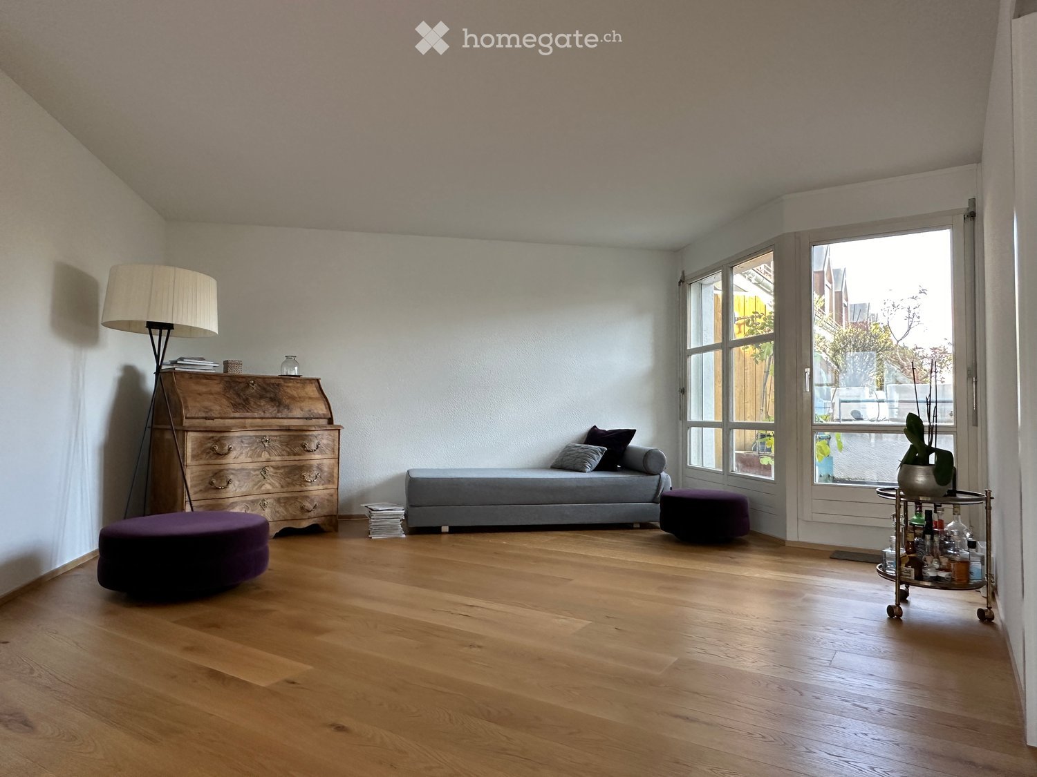 A modern living room with wooden flooring, a grey sofa, a purple ottoman, a wooden chest of drawers, a floor lamp, and a small cart with bottles and a vase.