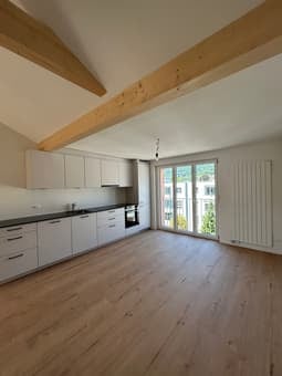 Bright, open kitchen with white cabinets, dark countertops, and large windows offering a view. The floor is made of light wood, and the space is completely empty with no furniture or appliances.