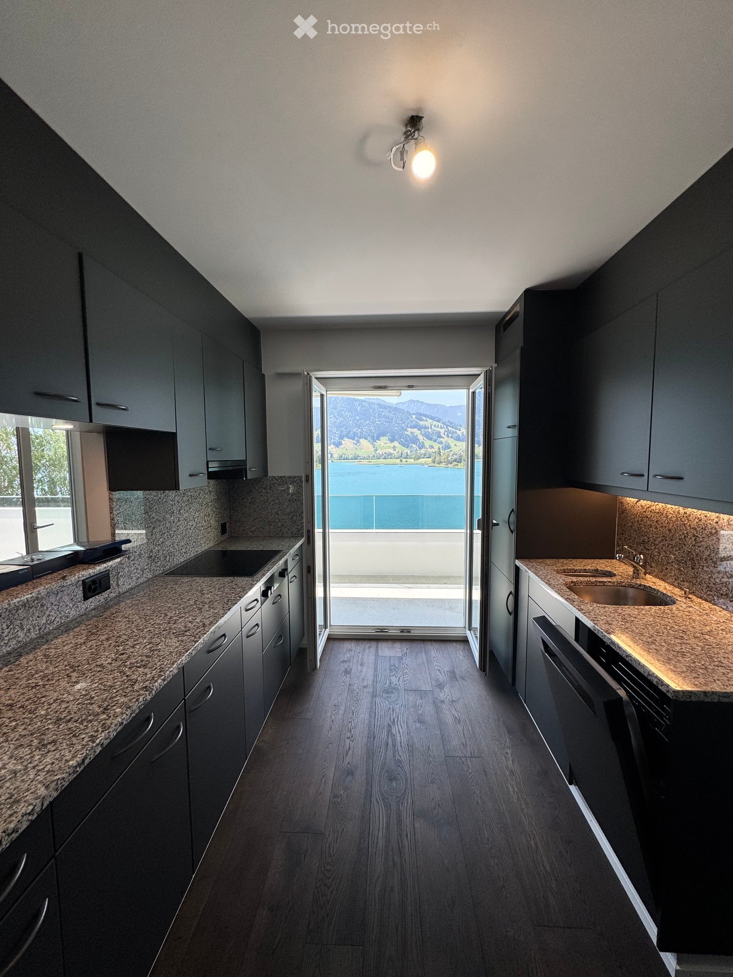 Modern kitchen with dark grey cabinets, granite countertops, and a view of a lake and mountains through a large window.