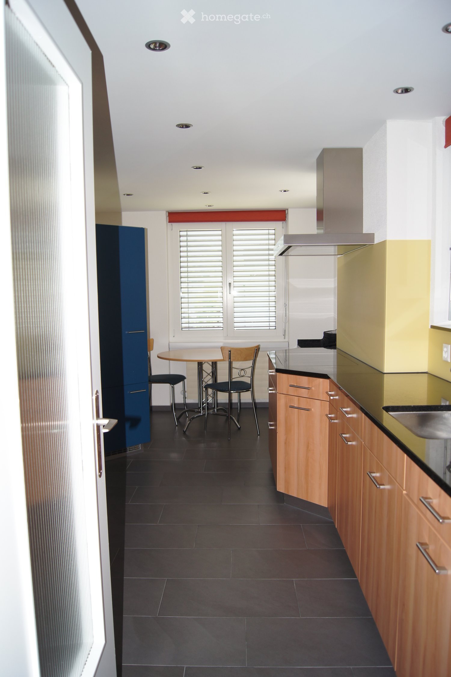 Modern kitchen with wooden cabinets, black countertops, a small round table with two chairs, and a window with blinds.