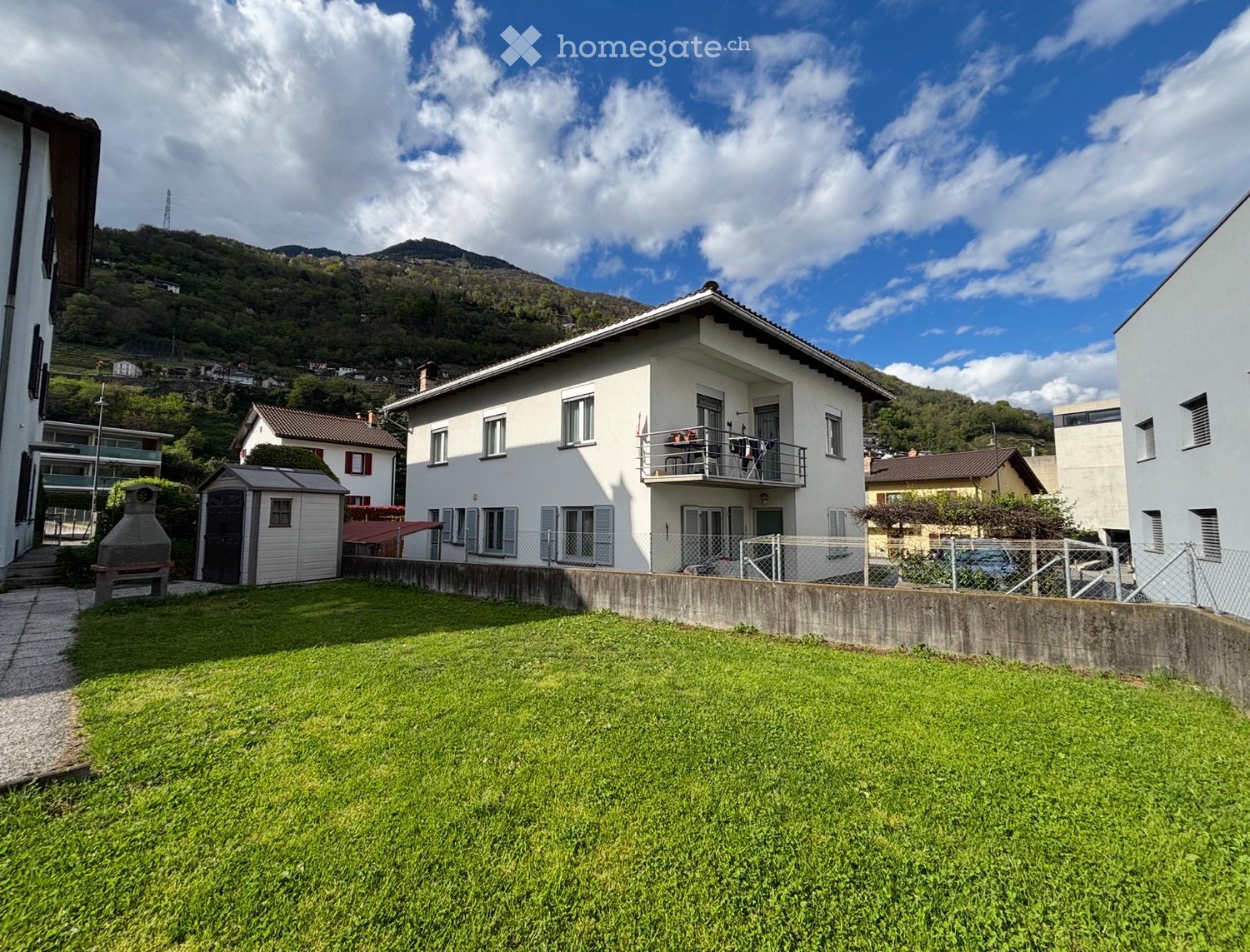 Two-story white house with multiple windows, a balcony, and a fenced yard with a small shed and barbecue area.