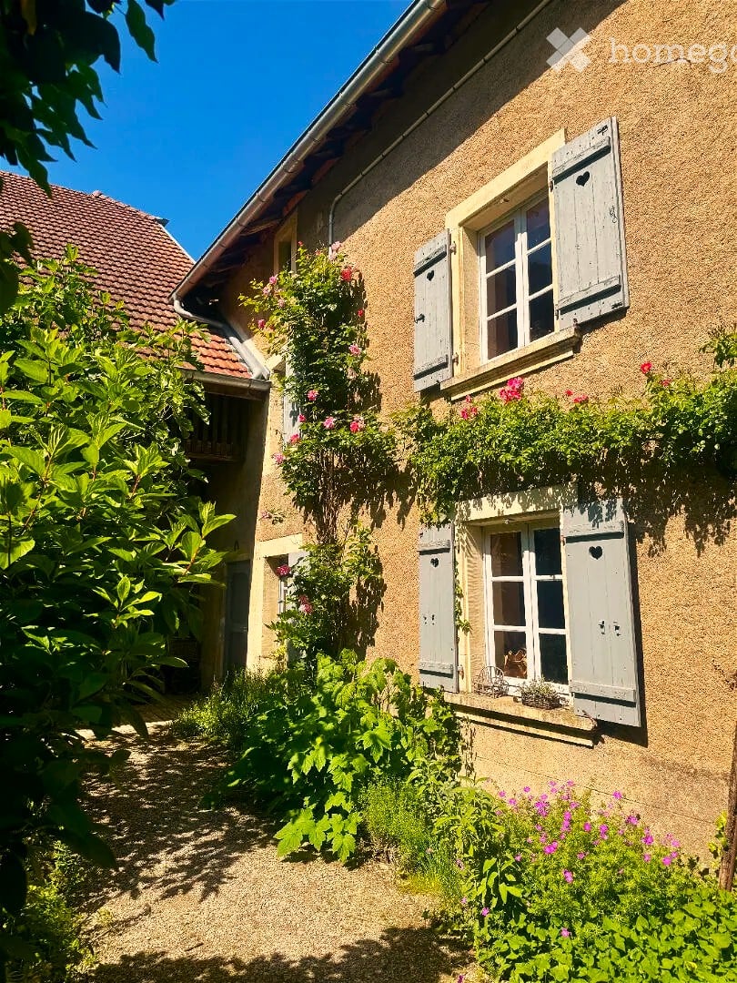 A charming, rustic building with beige stone walls, white shutters, and flower boxes, surrounded by lush greenery and a gravel pathway.