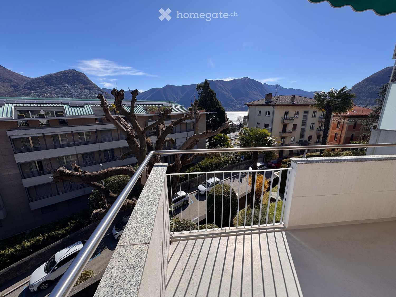 Balcony overlooking a scenic view of mountains, a lake, and surrounding buildings. The balcony has a metal railing and is part of a multi-story residential complex.