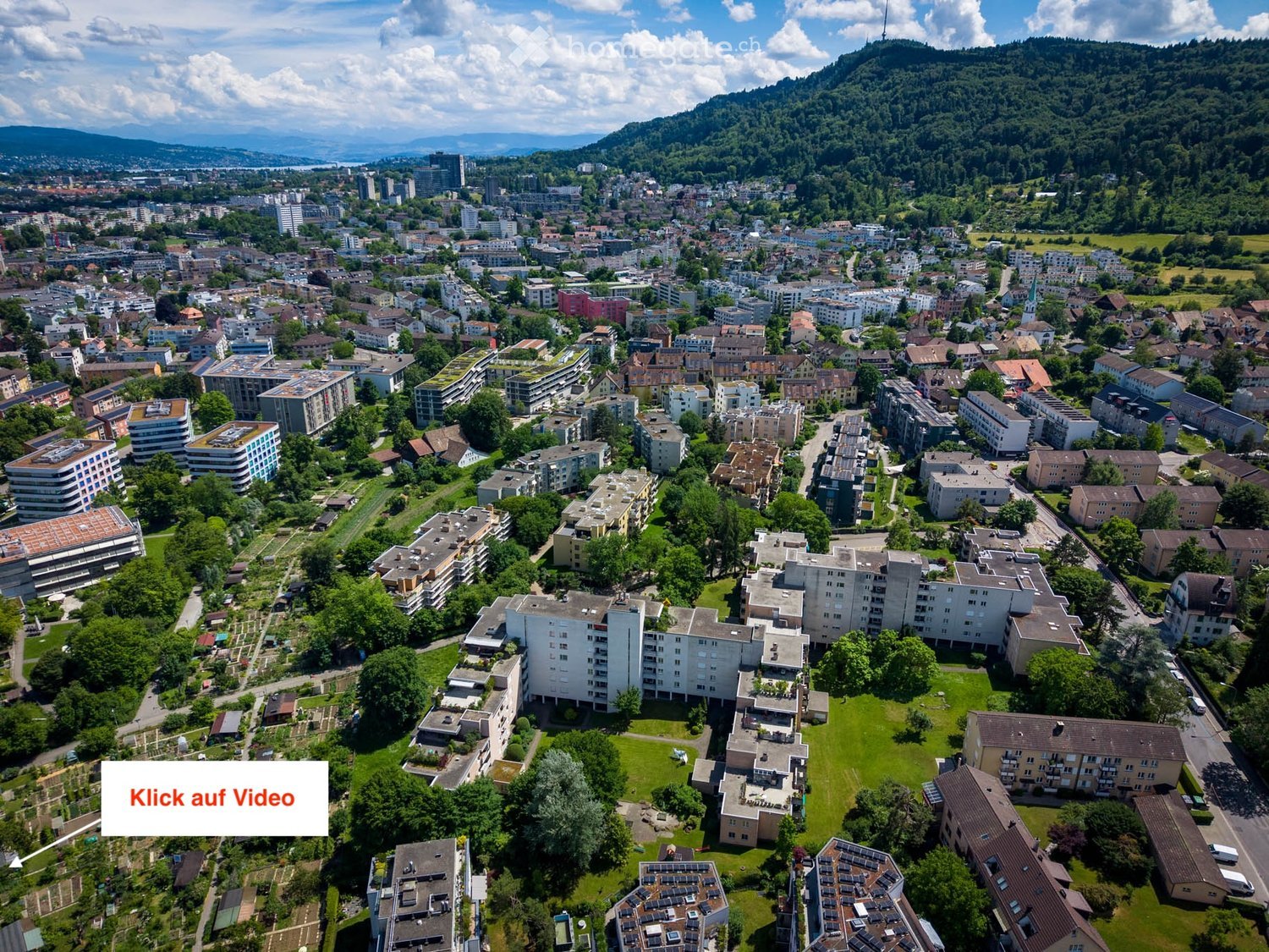 Aerial view of a cityscape with numerous residential and commercial buildings, surrounded by green spaces and trees, with distant mountains visible in the background.