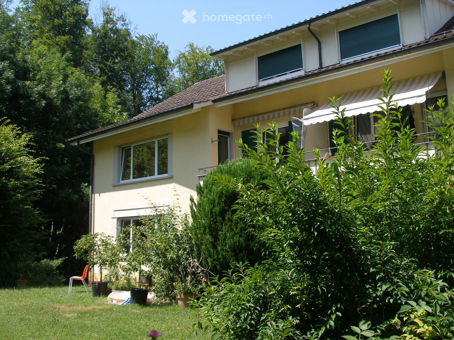 Two-story house with yellow siding, multiple windows, and a covered balcony area. The house is surrounded by a well-maintained garden with various plants and trees.