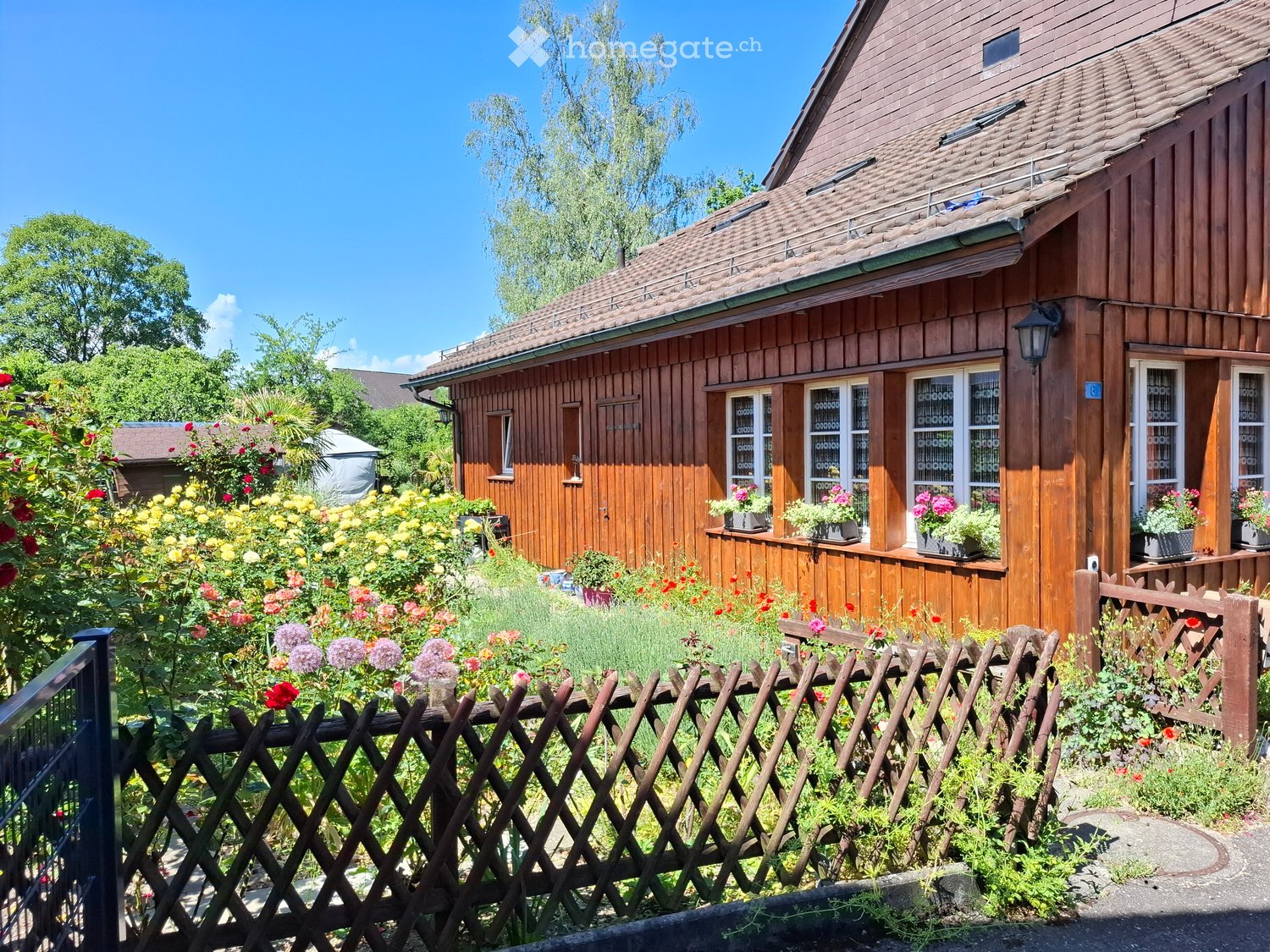 A wooden house with a sloped roof, surrounded by a garden with colorful flowers and a wooden fence.