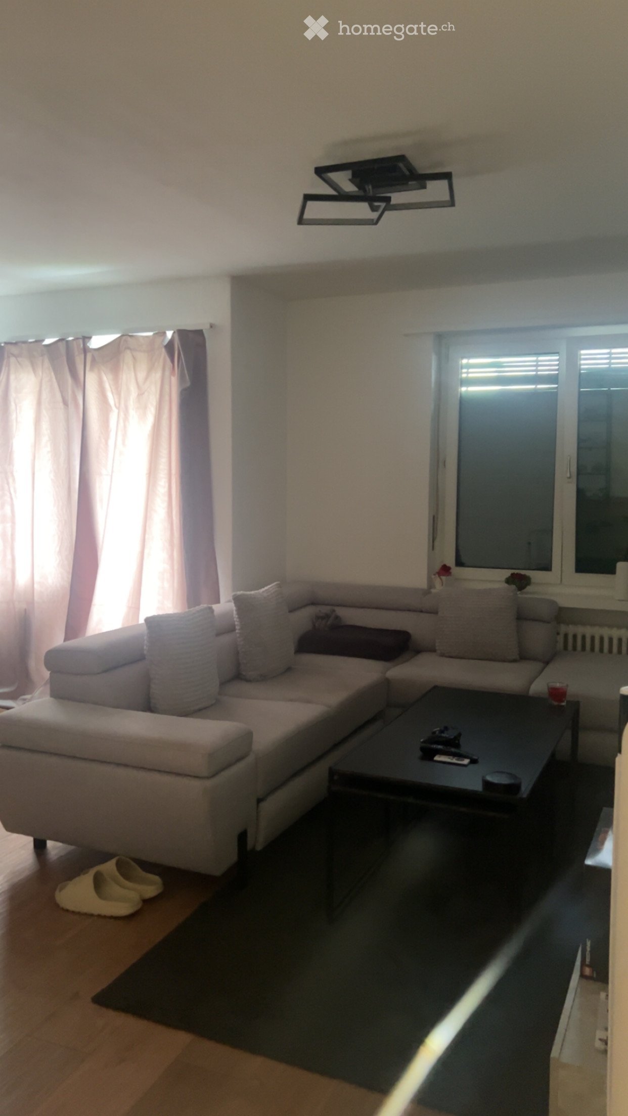 A modern living room with a grey sectional sofa, a black coffee table, and a pair of beige slippers near the entrance. The room features large windows with curtains and a sleek ceiling light fixture.