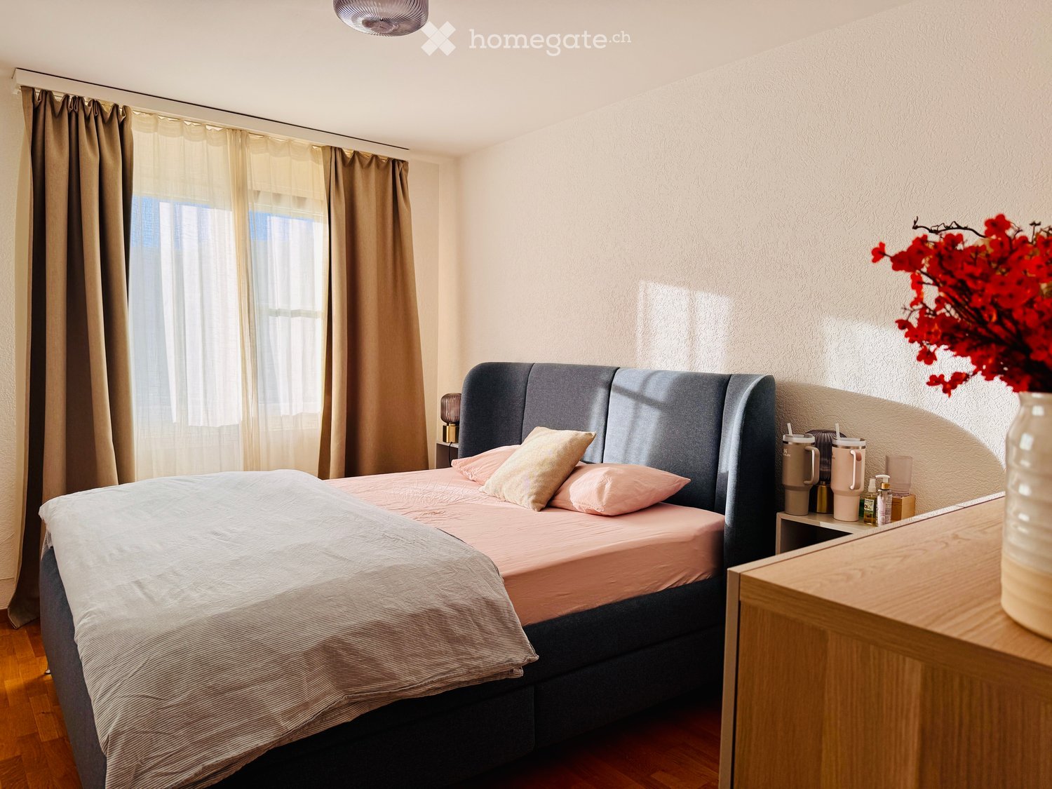 A modern bedroom with a large bed, grey headboard, pink bedding, nightstand, and decorative vase with red flowers on a wooden side table.