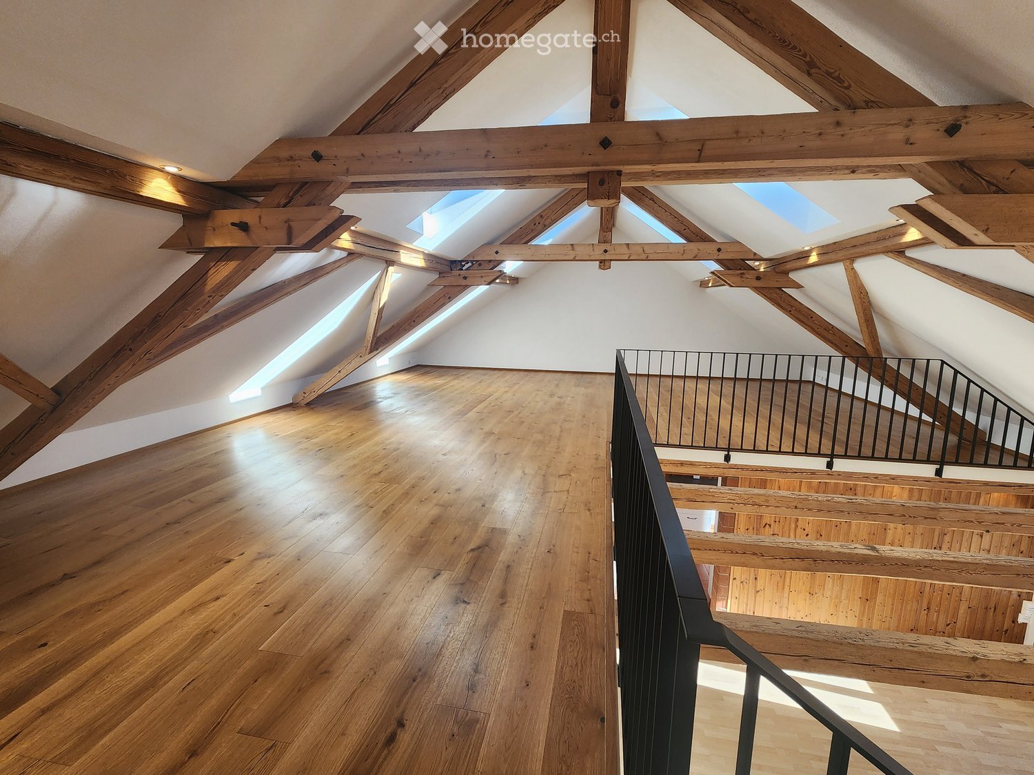 An empty attic space with exposed wooden beams, skylights, and wooden flooring, featuring a staircase leading downwards.