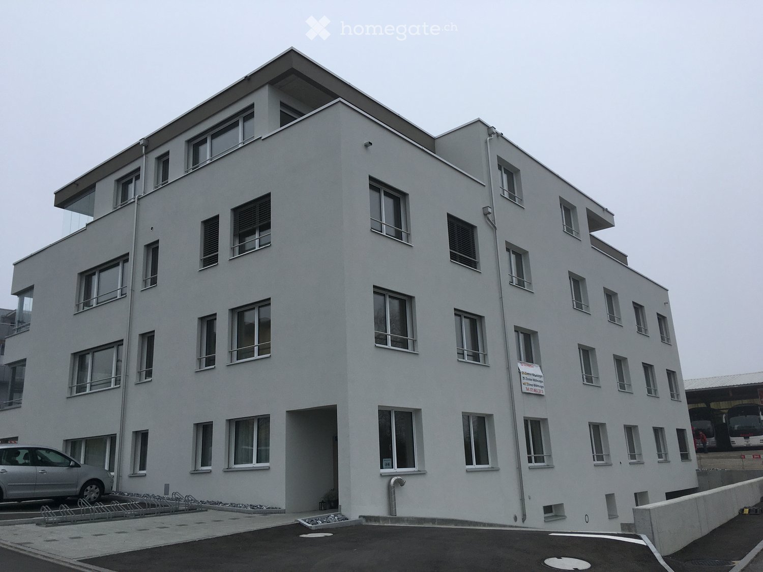 Modern multi-story building with white facade, numerous windows, and balconies. Features a parking area with a car and bicycles.
