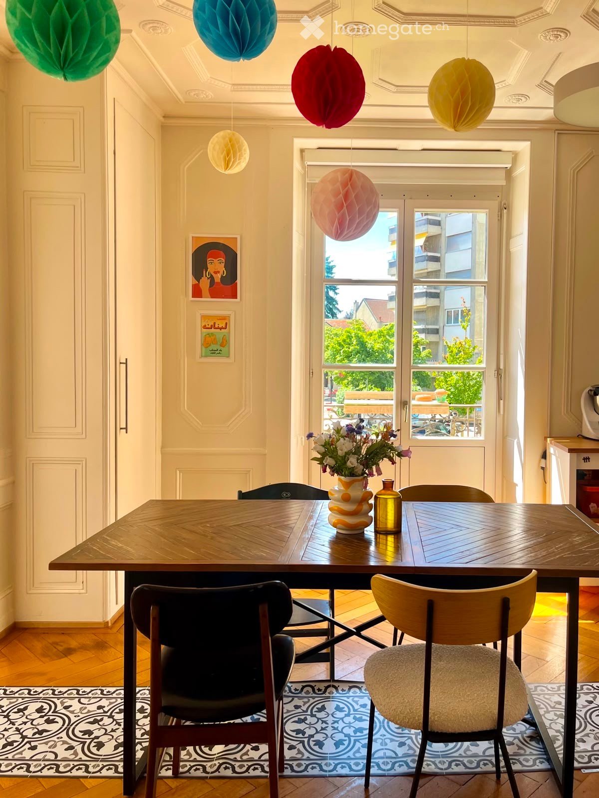 Dining area with wooden table, chairs, vase with flowers, and decorative items. Large windows provide natural light and a view of the street.