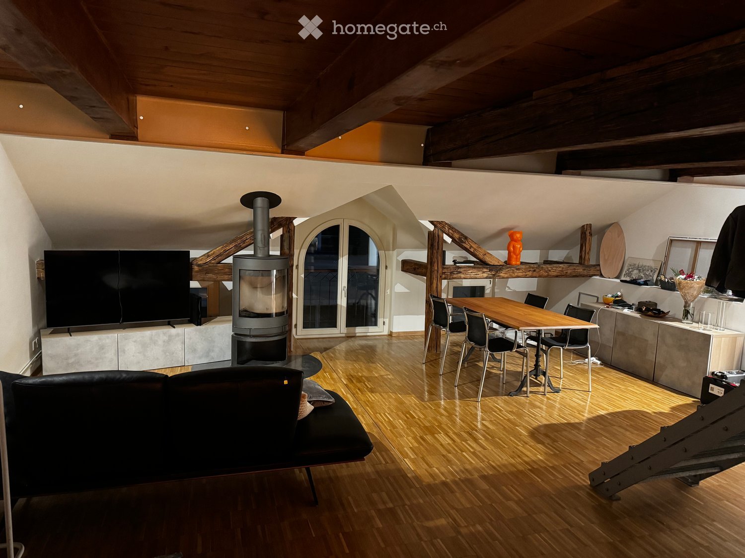Open-concept living and dining area with wooden flooring, exposed wooden beams, a modern fireplace, a large TV, a dining table with chairs, and a kitchen counter.