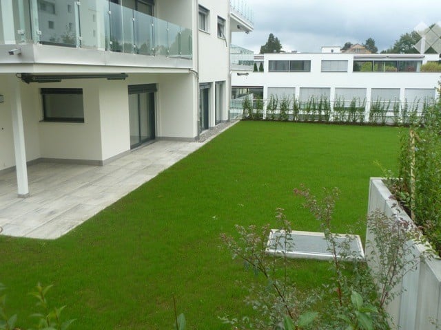 Modern outdoor garden area with artificial grass, surrounded by white building walls and glass balcony railings. Features include a paved patio area and small raised planters.