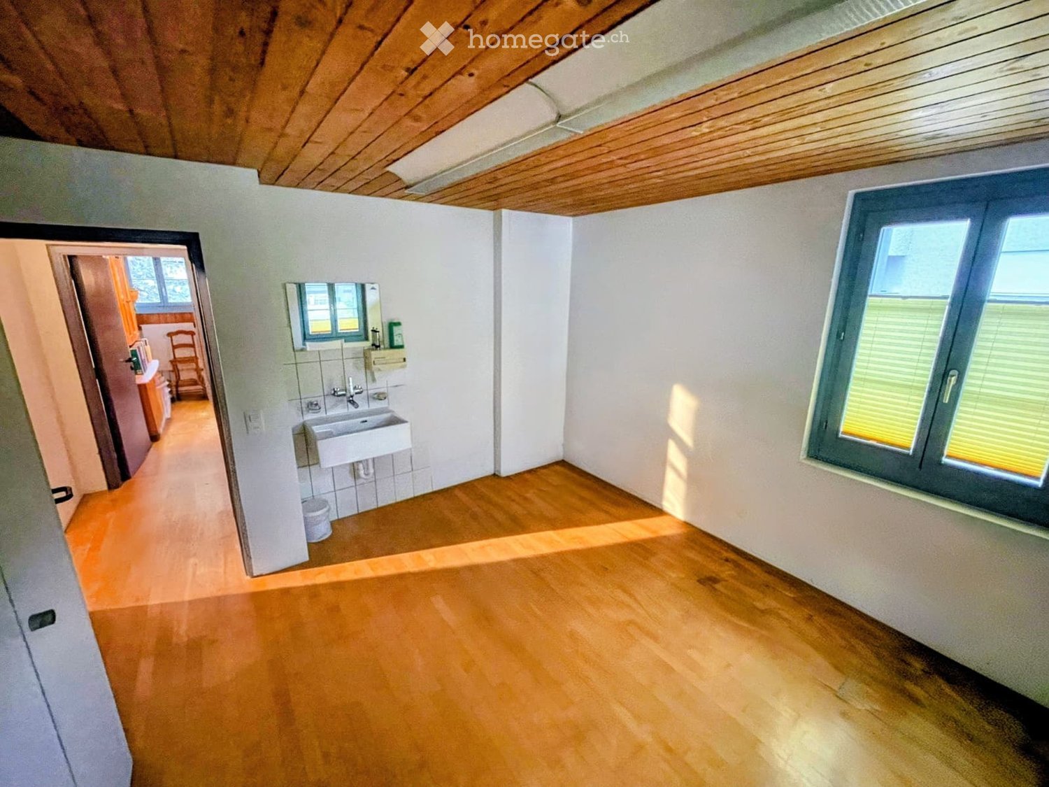 Empty room with wooden flooring, white walls, and a large window with blinds. A doorway leads to another room with a sink and mirror visible.