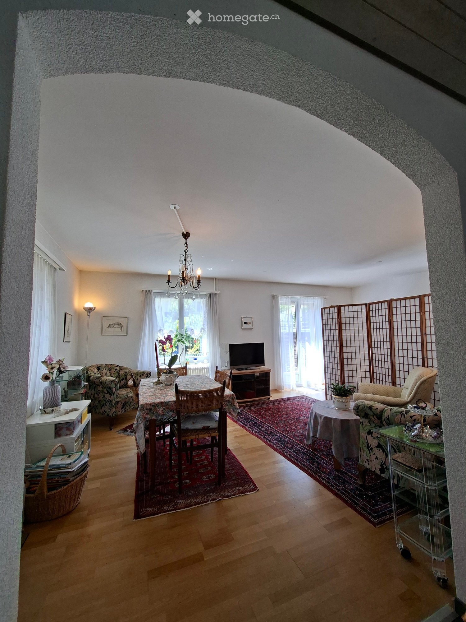 Spacious living and dining area with wooden flooring, large windows, and high ceilings. Features a sofa, dining table with chairs, television, chandelier, and decorative items.