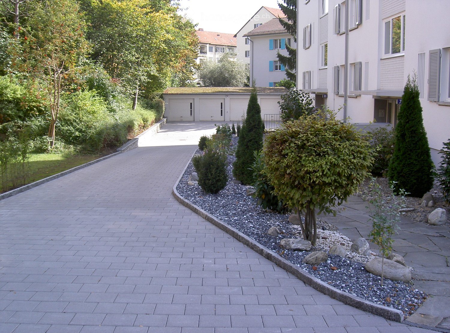Brick driveway leading to a garage with surrounding greenery and decorative rock garden.