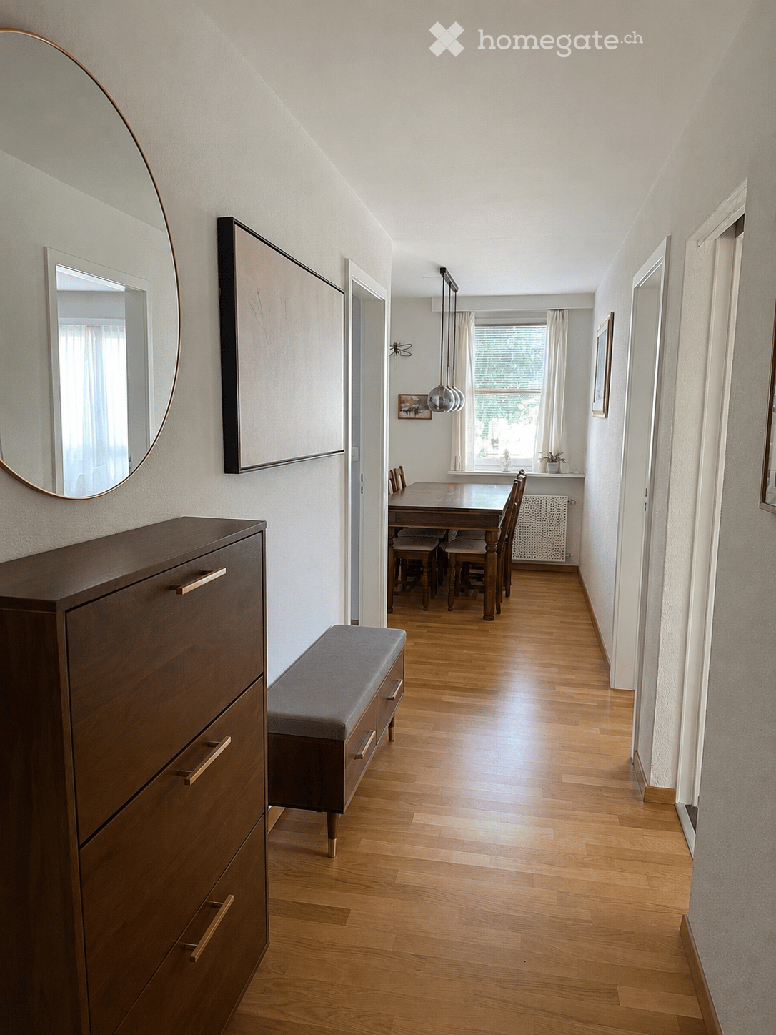A well-lit hallway with wooden flooring leads to a dining area featuring a wooden dining table and chairs, a grey bench, and a wooden cabinet with brass handles. A large round mirror and picture frame adorn the walls, and pendant lights hang from the ceil