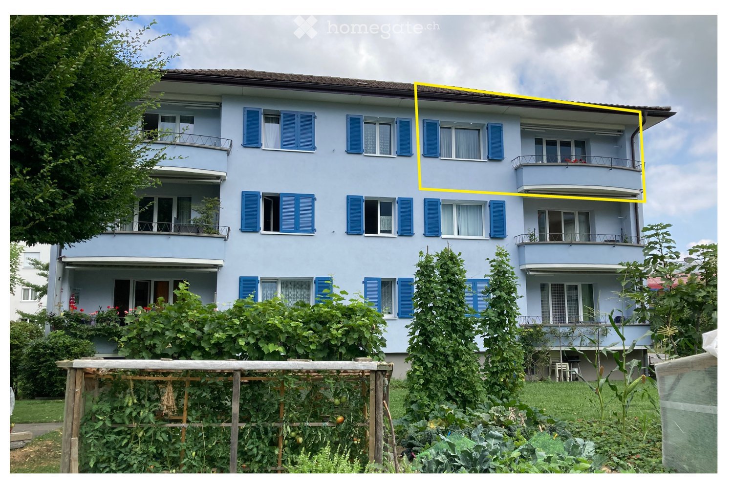 A three-story residential building with light blue walls, blue shutters, and multiple balconies. The building is surrounded by lush greenery and a garden area with vegetables and plants.