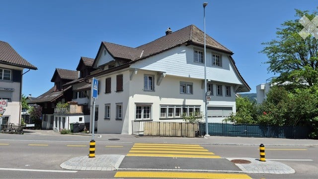 A multi-story residential building with white siding and dark brown roof, featuring multiple windows and a small balcony area.