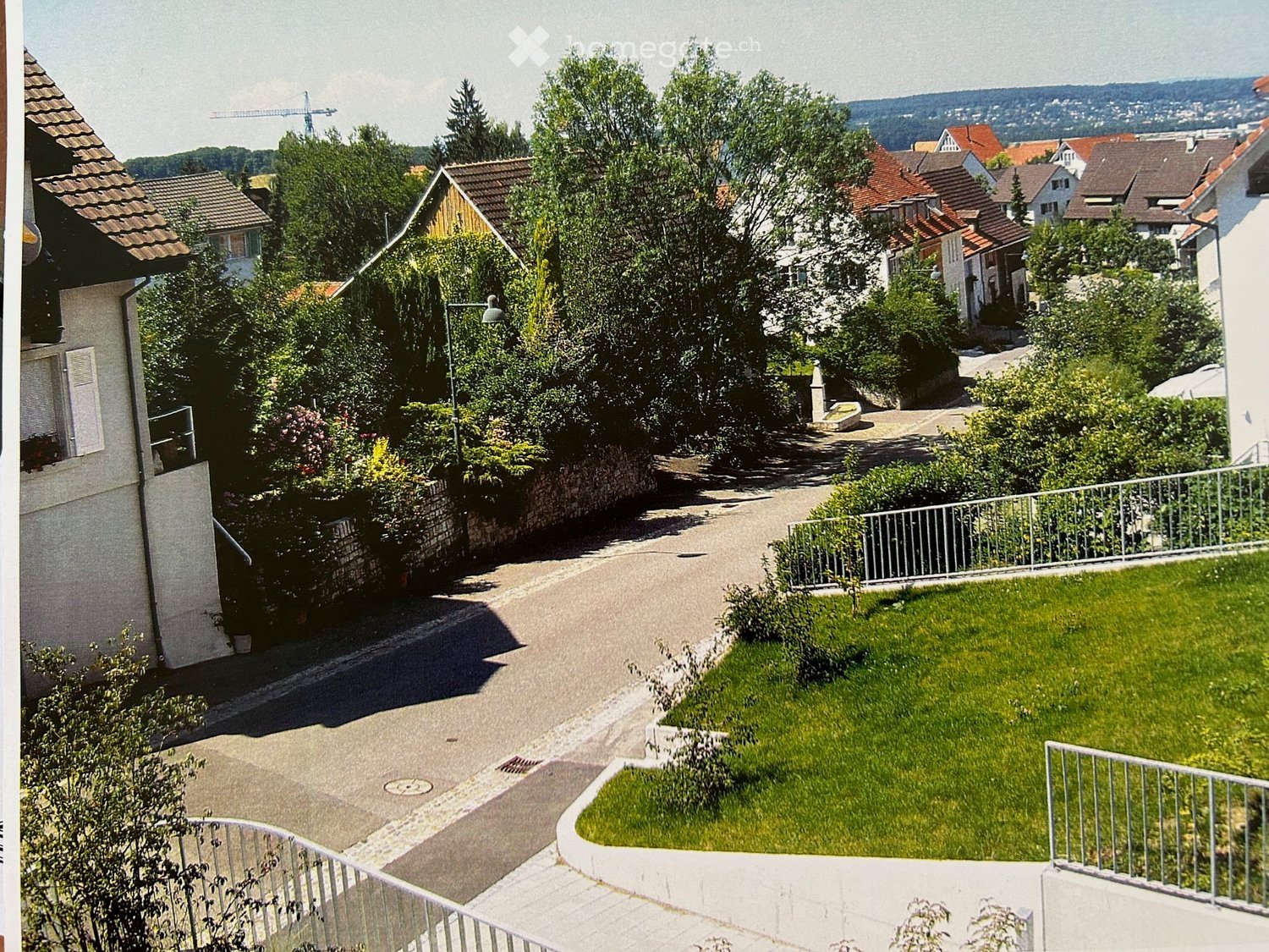 A street view of a neighborhood with houses, trees, and a crane in the distance. The houses have brown roofs and white walls. The street is lined with trees and bushes, and there is a metal fence on the right side. The sky is clear and blue.
