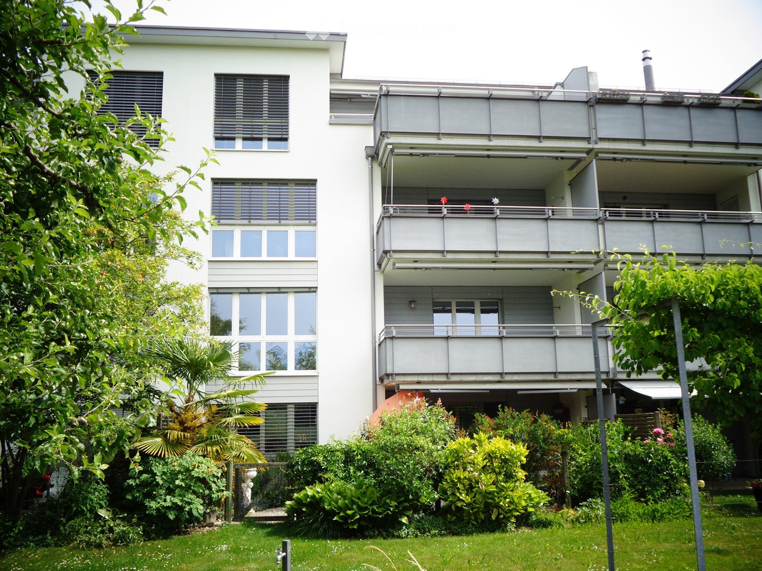 Modern two-story building with white facade, large windows, and balconies. Surrounded by lush greenery and a well-maintained lawn.