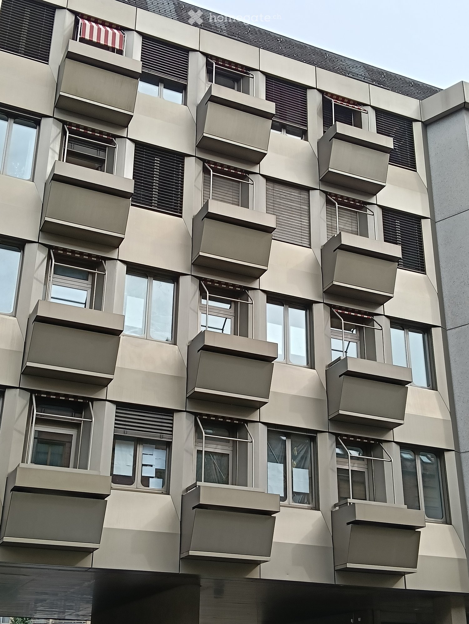 Modern multi-story building with numerous balconies and large windows, featuring a clean, geometric design with beige and grey tones.