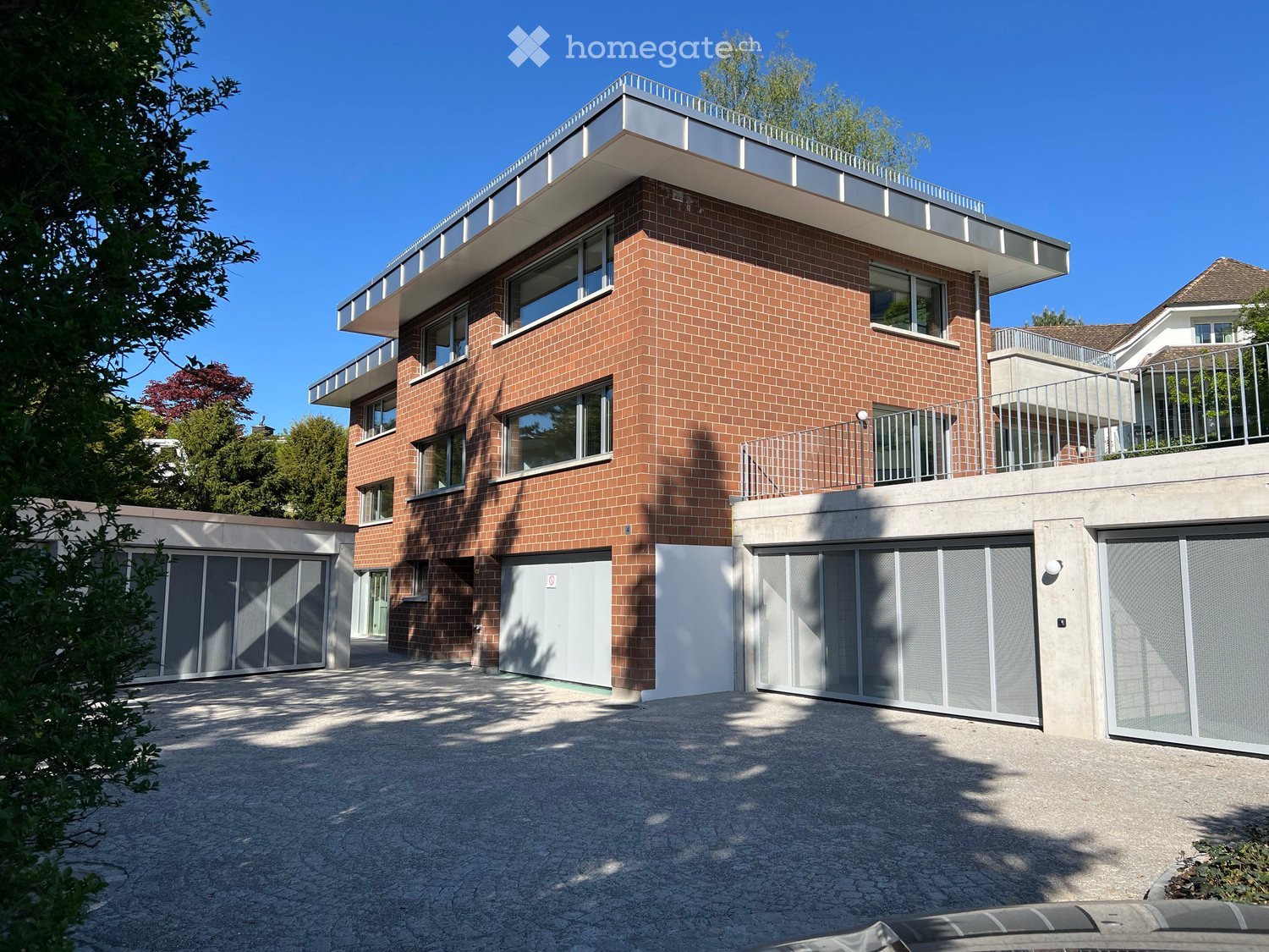 Modern two-story house with red brick facade, large windows, and multiple balconies. Features include a spacious driveway, attached garage, and contemporary design.