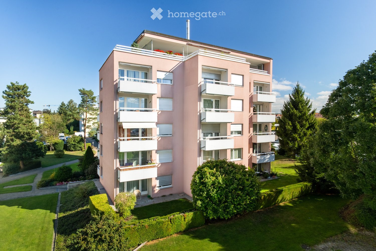 A multi-story apartment building with a pink exterior and white balconies. The building is surrounded by lush greenery and landscaping, including trees and shrubs.