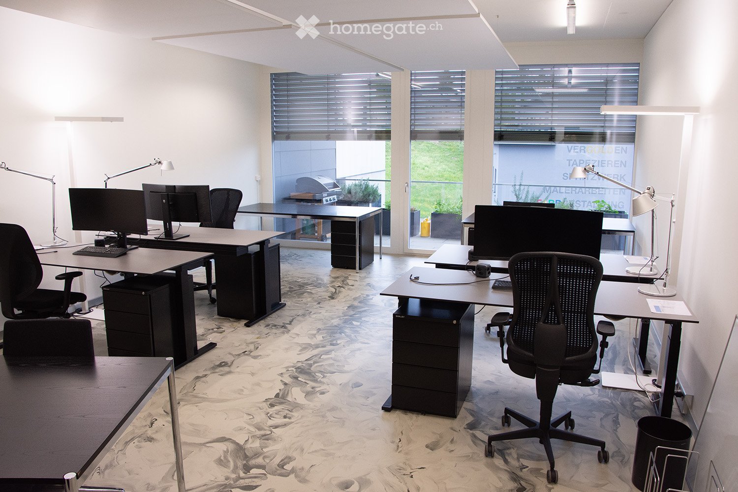 Modern office space with multiple desks, ergonomic chairs, computers, keyboards, and lamps. Large windows with blinds provide natural light.