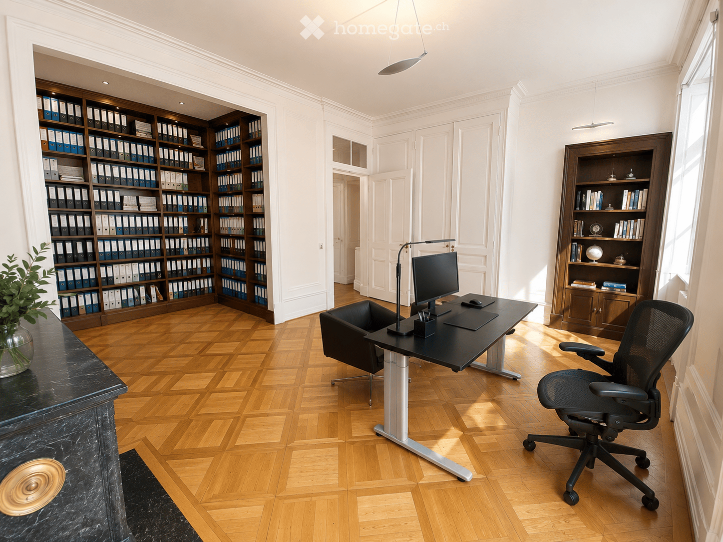 Elegant office with wooden herringbone flooring, large black desk, ergonomic chair, computer monitor, bookshelf filled with binders and books, and a decorative vase with greenery.