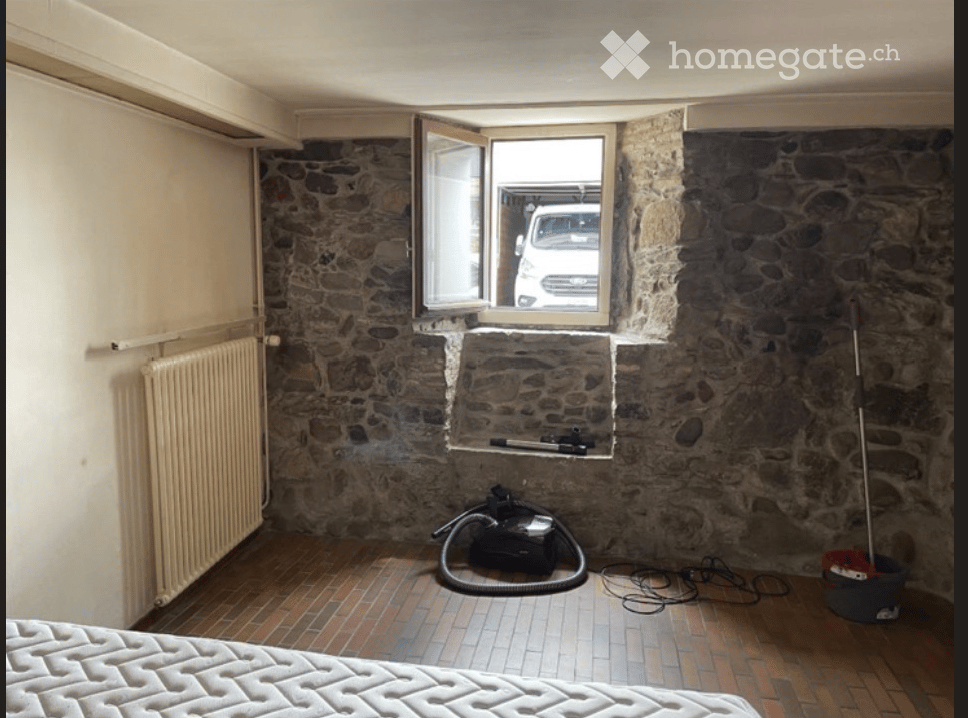 A small, rustic room with exposed stone walls, a tiled floor, and a single bed. A window with an open shutter reveals a parked vehicle outside. Cleaning supplies and a vacuum cleaner are visible on the floor.