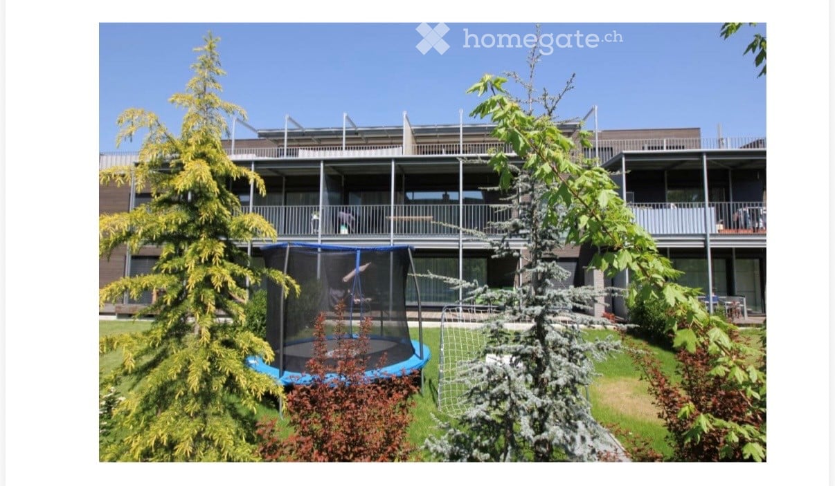 A modern multi-story building with balconies, surrounded by a well-maintained garden featuring a trampoline, trees, and shrubs.