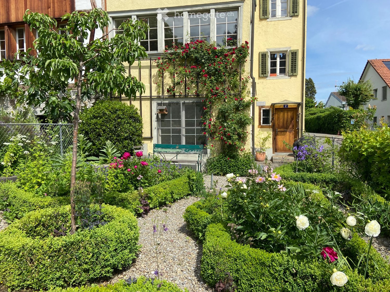 Well-maintained garden with neatly trimmed hedges, colorful flowers, a gravel pathway, and a small bench near the house entrance.