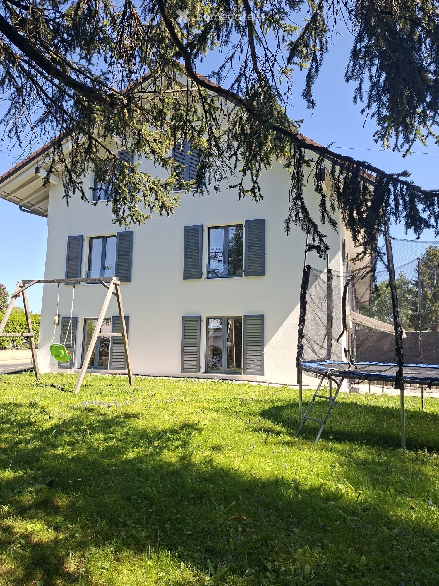 A two-story white house with multiple windows and shutters, surrounded by a grassy yard with a swing set and a trampoline.