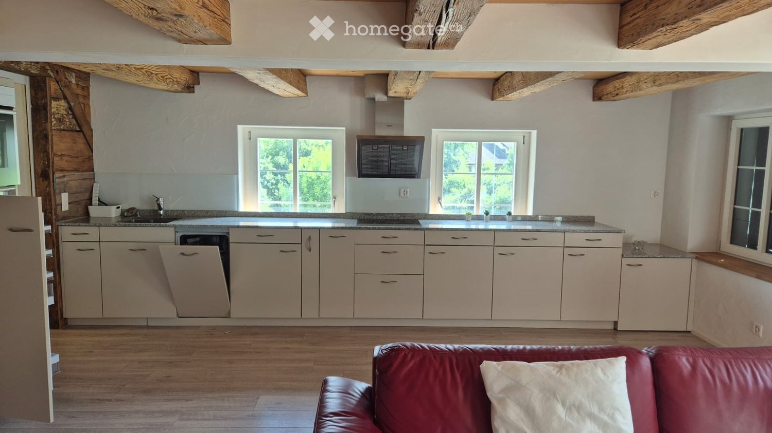 Open-plan kitchen and living area with light-colored cabinets, a red leather sofa, and wooden flooring.