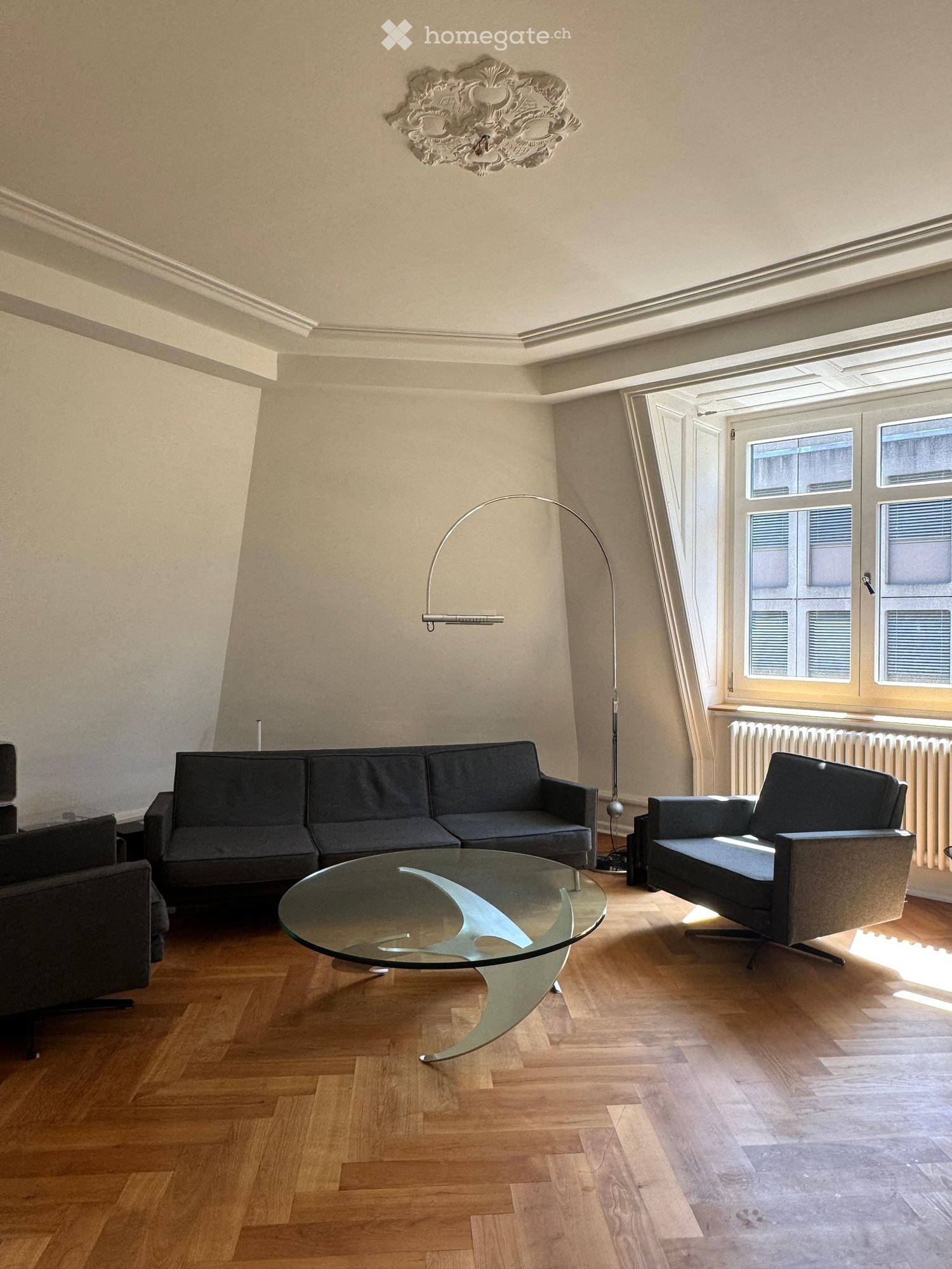 Modern living room with wooden flooring, a dark grey sofa, a glass coffee table, a floor lamp, and a decorative wall piece.