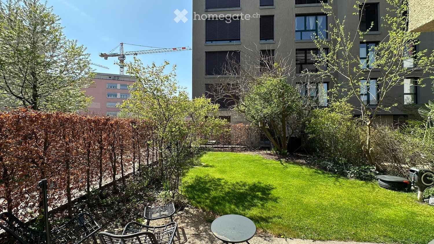 small garden with green grass, trees, shrubs, chairs, table, and a building in the background