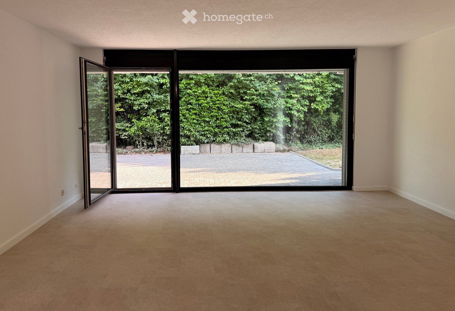 Empty room with light-colored walls and carpeted floor, featuring large sliding glass doors that open to a view of greenery and a paved area.