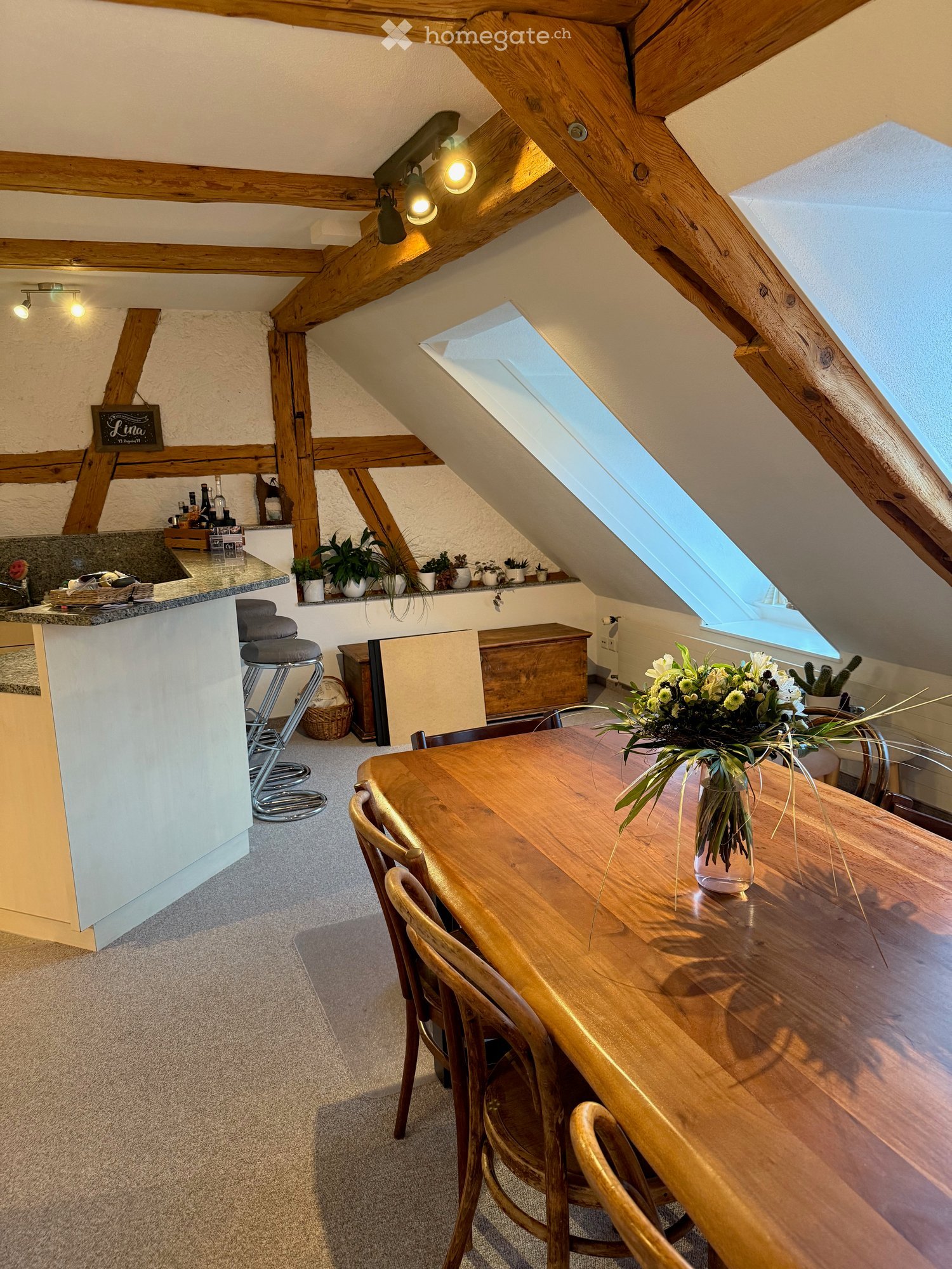 Cozy attic space with wooden beams, skylight windows, dining table with chairs, bar area with stools, and decorative plants.