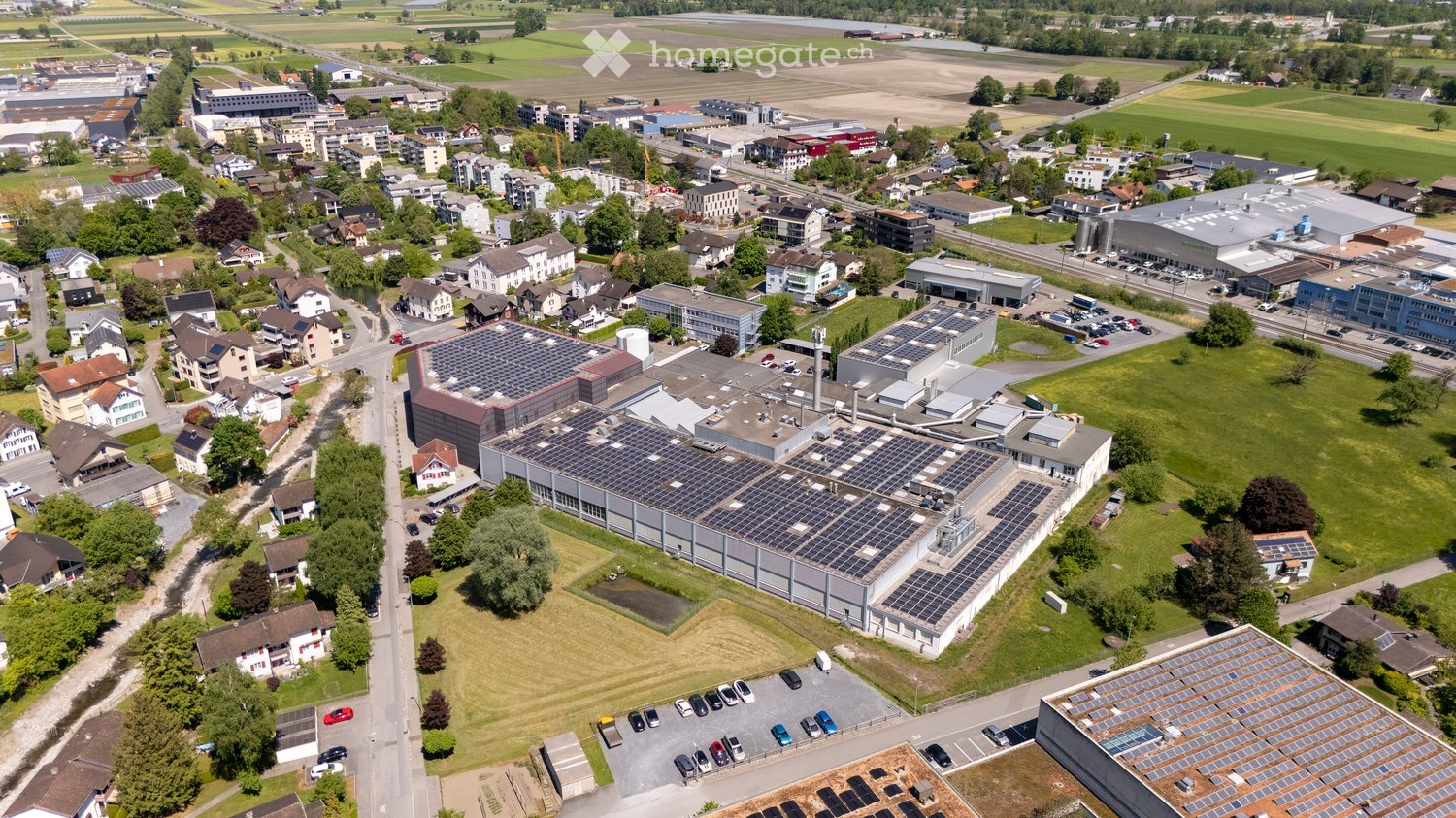 Aerial view of a suburban area with residential houses, industrial buildings, and green fields. Several buildings have solar panels on their roofs.