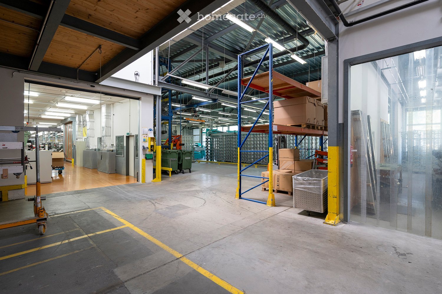 Large industrial warehouse with metal shelving units, cardboard boxes, and various equipment. The space features a high ceiling with exposed beams and fluorescent lighting, along with a concrete floor marked with yellow lines.