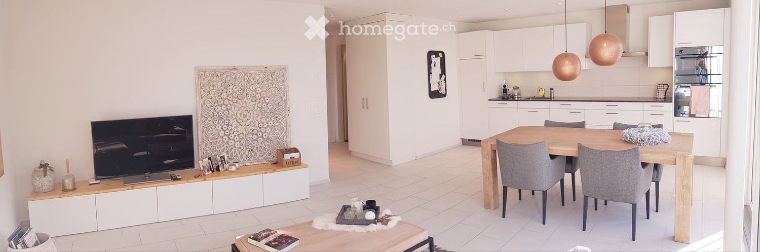 Modern open-plan living and dining area with white walls, tiled flooring, a TV stand, a dining table with chairs, and a kitchen area with cabinets and appliances.