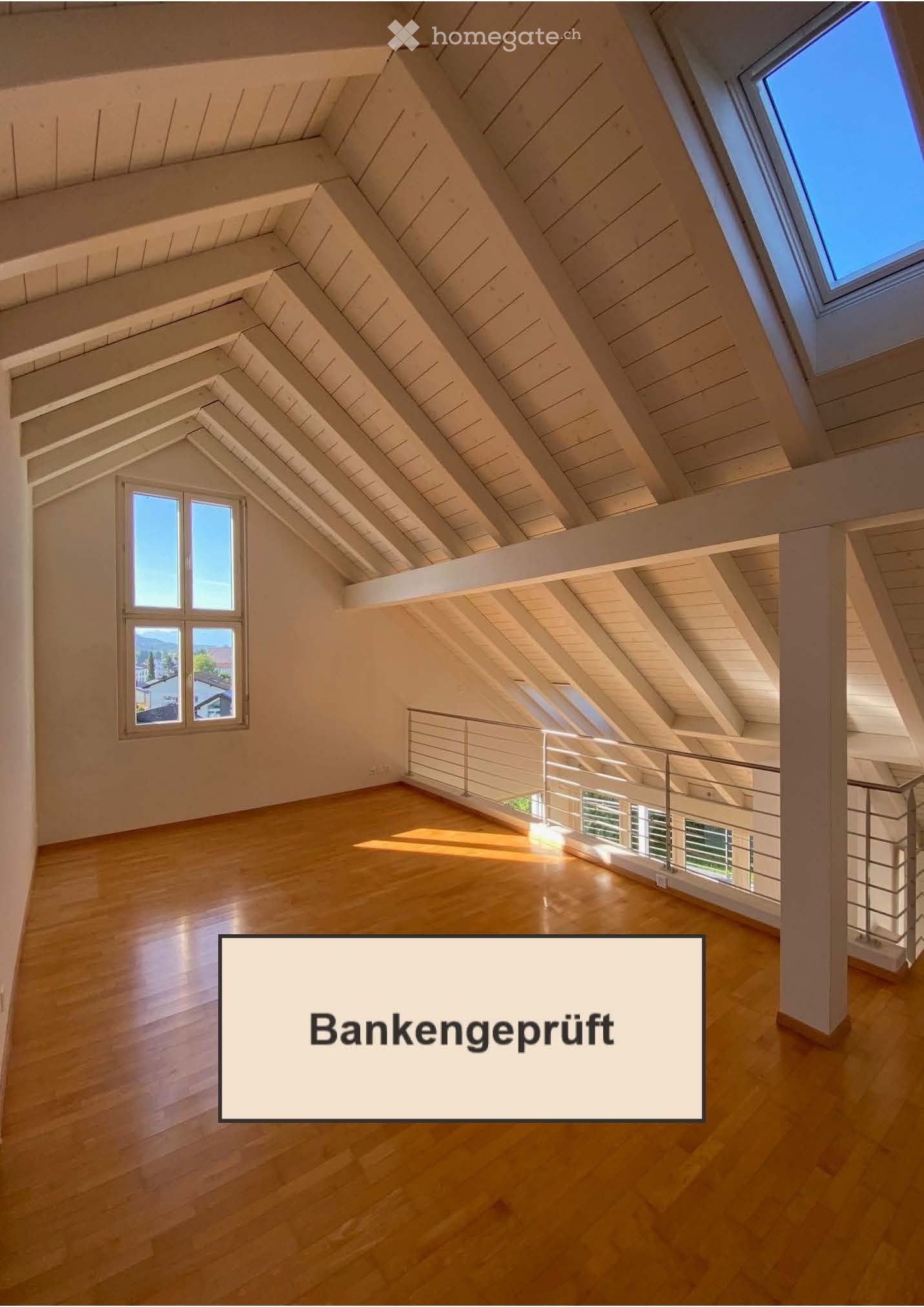 Bright, empty attic space with wooden floors, exposed wooden beams, large windows, and a modern metal railing overlooking a lower level.