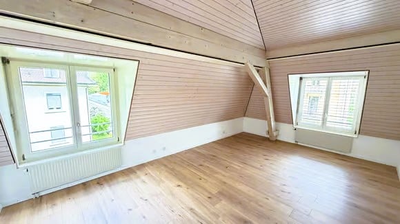 Empty room with wooden flooring, white walls, and large windows with white frames. The room has a slanted ceiling with exposed wooden beams.