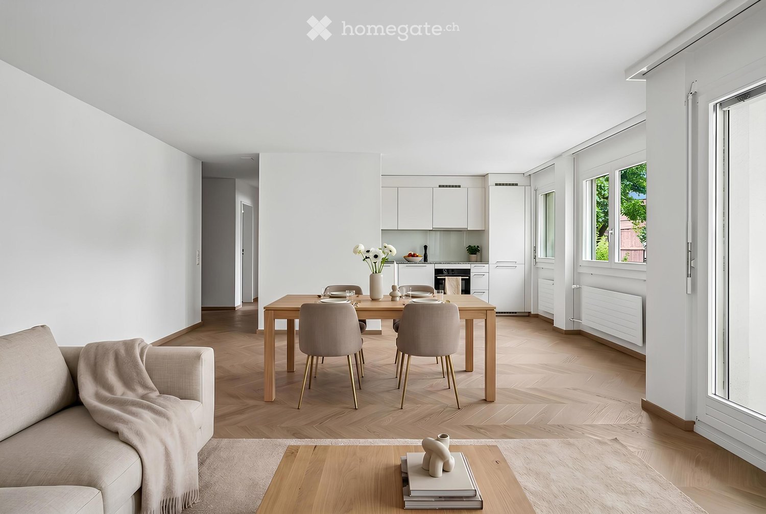 Bright, open-plan living and dining area with light wood flooring, white walls, and large windows. Features a sofa with a throw, coffee table with decorative items, dining table with chairs, and a modern kitchen area with white cabinets and stainless stee